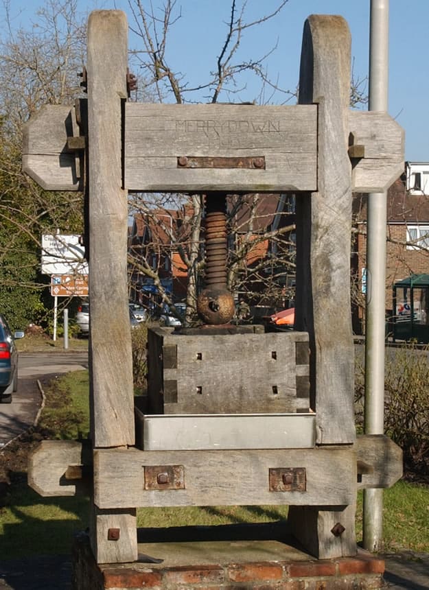 A large wooden cider press stands outdoors, with a metal screw mechanism in the center. The word "MERRYDOWN" is carved on the top beam. Trees, a path, and parked cars are visible in the background.
