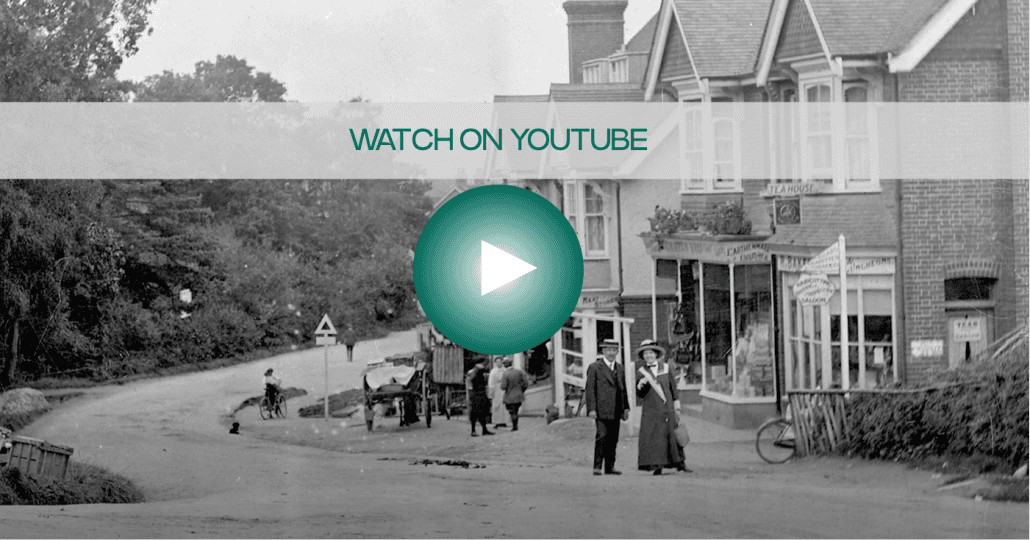 A sepia-toned image of an old village street with people standing near buildings. A large, transparent "Watch on YouTube" overlay with a play button is centered. Trees and a road are visible in the background.