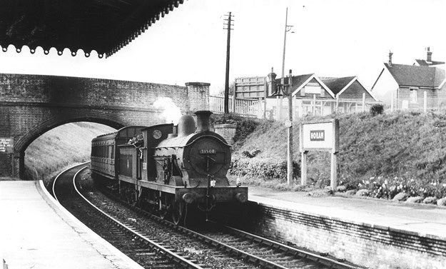 A steam train approaches a platform at a rural station. The station name sign reads "WOLAN." A bridge arches over the tracks, with small buildings and telephone poles visible in the background. The scene is black and white, suggesting a historical time.